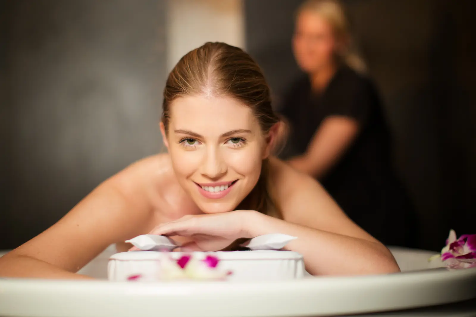 Lächelnde Frau in einer Badewanne mit Blüten, im Hintergrund eine Therapeutin